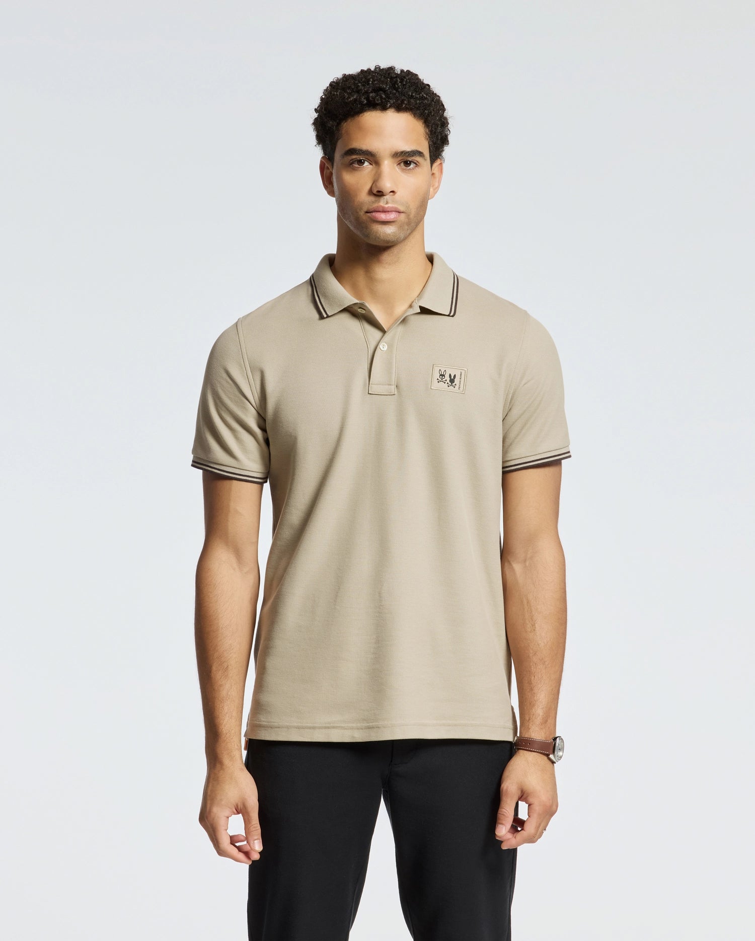 A man is wearing the Psycho Bunny MENS MONROE PIQUE POLO in a regular fit beige Pima cotton, made in Peru, featuring a small emblem on the chest. He is also wearing black pants and standing against a plain white background.