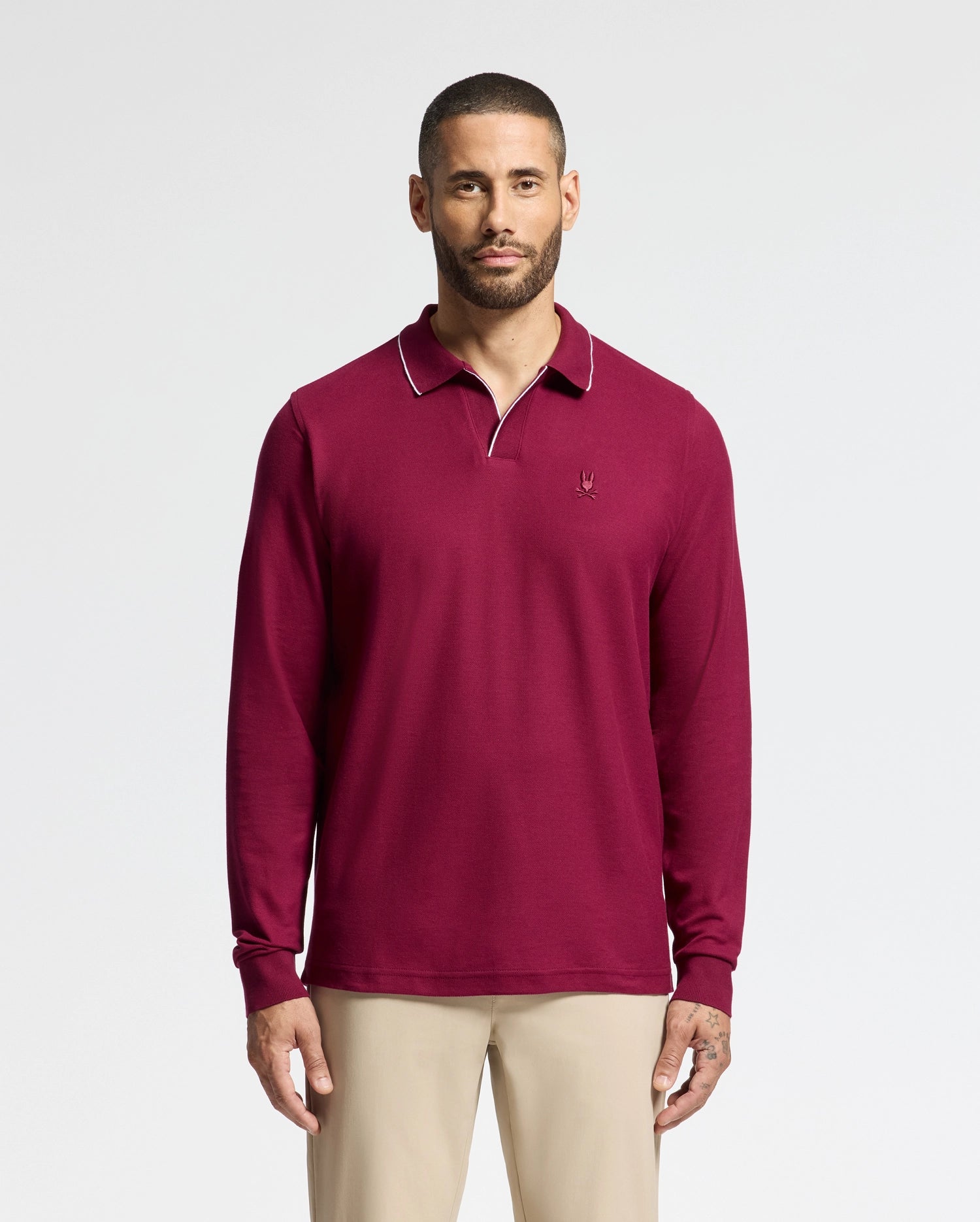 A person wearing a Psycho Bunny MENS CREEDE PIQUE JOHNNY COLLAR POLO - B6M265E200 in maroon stands against a plain white background. They have short hair and a beard. The shirt, which has a small embroidered logo on the chest, is paired with light-colored pants.