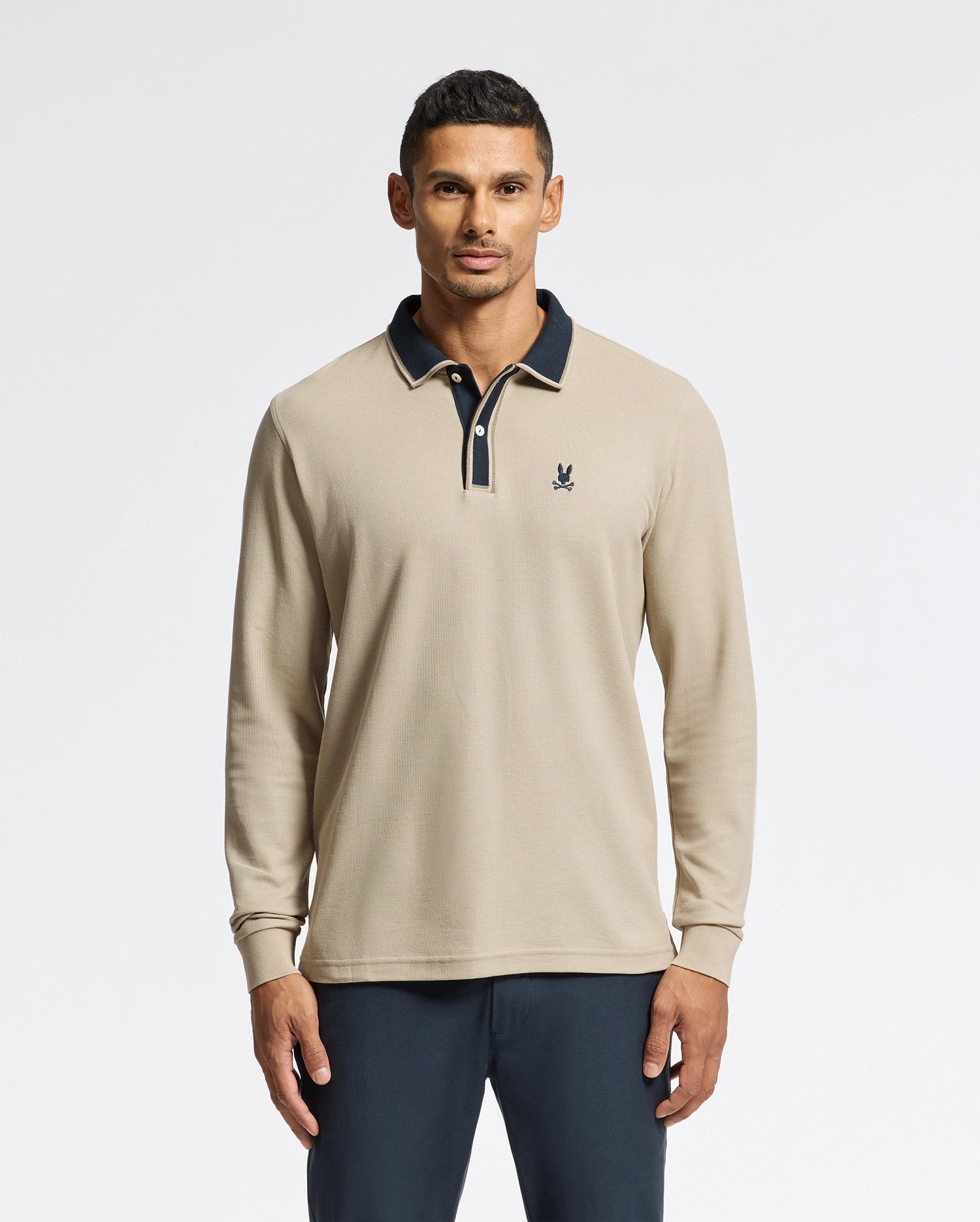 A man stands against a plain white background, showcasing the Psycho Bunny MENS KENT LONG SLEEVE PIQUE POLO in beige Pima cotton, featuring a dark collar and cuffs. Paired with dark pants, this ensemble exudes quiet luxury, highlighted by the small logo on the left side of the shirt.