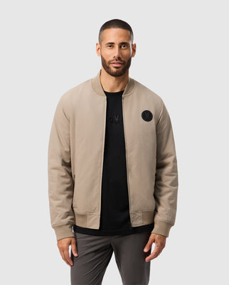 A man with short hair and a beard models the Psycho Bunny MENS SPRUCE BOMBER JACKET - B6N804U1CO in beige over a black shirt. He stands against a plain light gray background, looking directly at the camera.