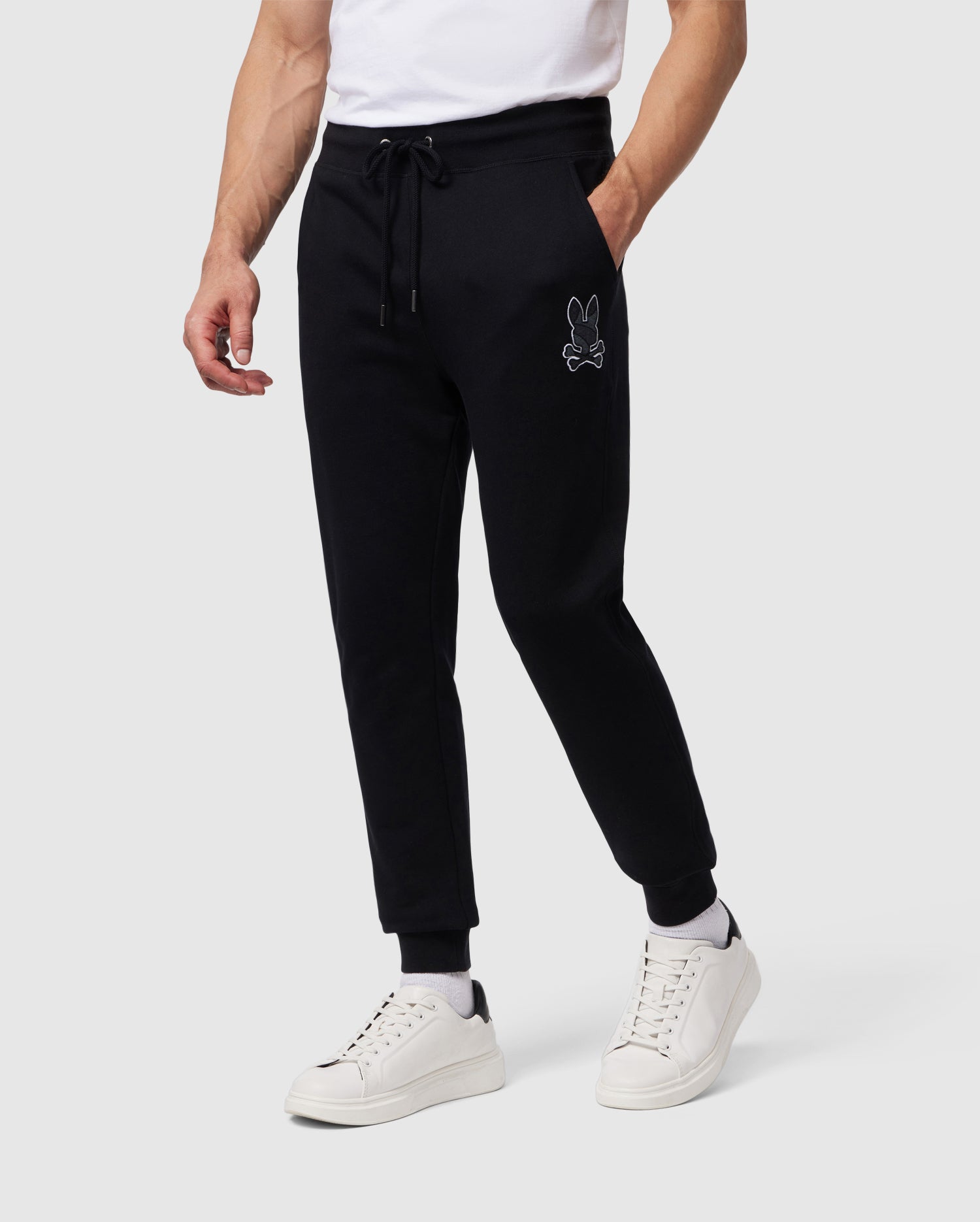 A person dressed in black Psycho Bunny MENS LENOX REGULAR FIT SWEATPANT - B6P105B200 with a chainstitch-embroidered bunny logo on the left thigh and a white t-shirt. They are also wearing white sneakers. The background is plain and neutral.