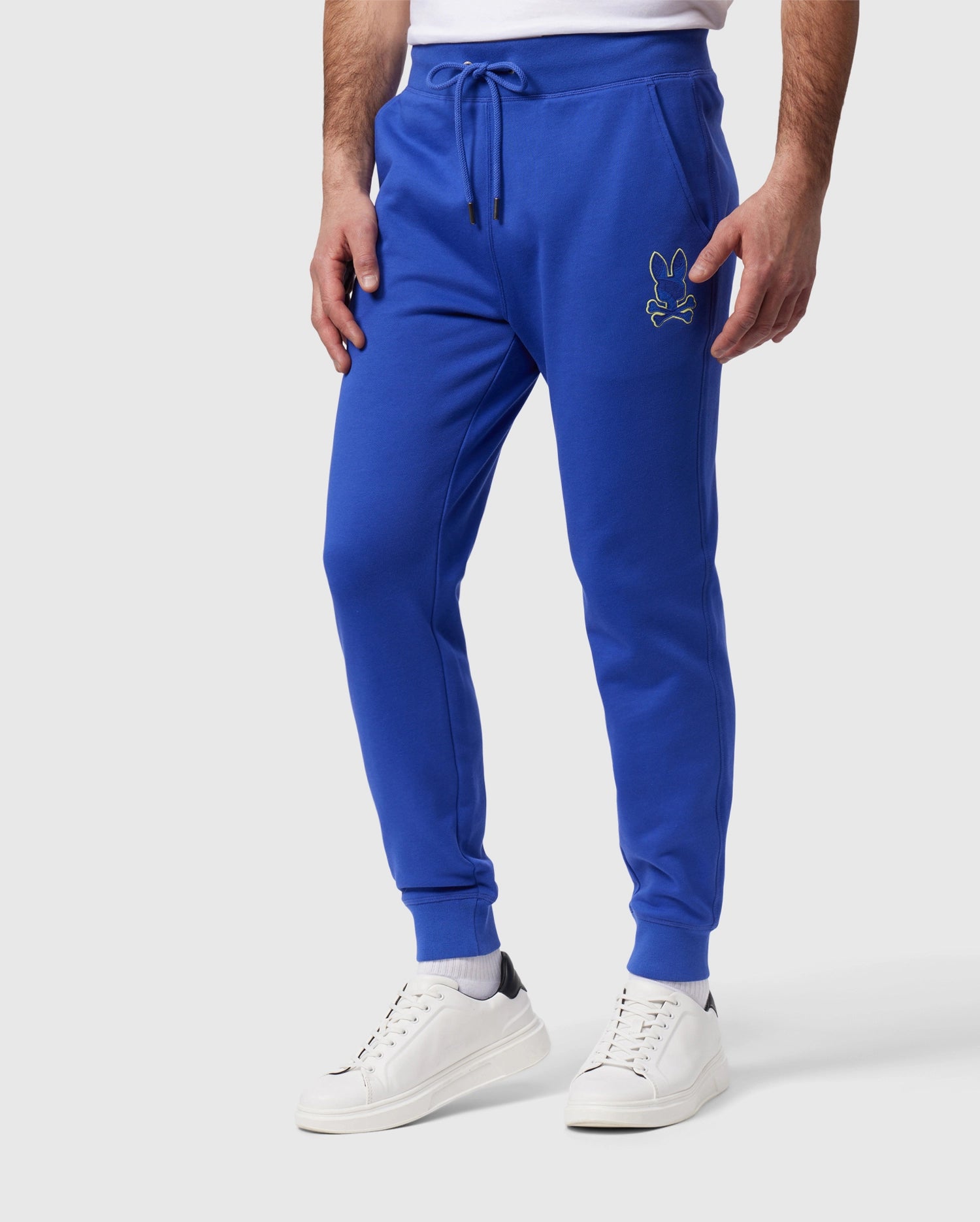A person is wearing bright blue MENS LENOX REGULAR FIT SWEATPANT - B6P105B200 by Psycho Bunny made from cotton fleece, featuring an embroidered cartoon character on the upper left thigh. The pants have a drawstring waist and cuffs at the ankles. The person is also sporting white sneakers with black accents on the heel. The background is plain white.