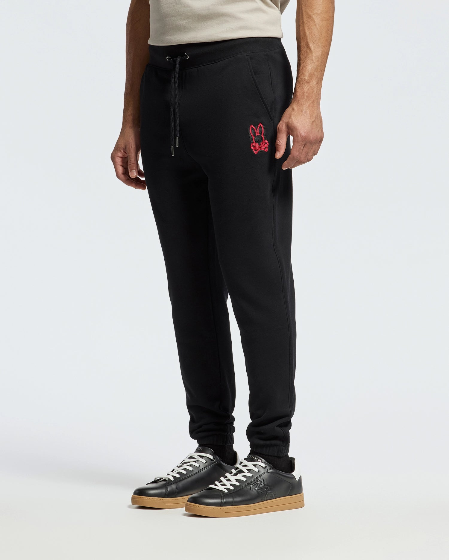 A person wearing the MENS PIERCE ESSENTIAL FRENCH TERRY SWEATPANT by Psycho Bunny, featuring a red bunny logo on the left side, paired with black sneakers that have tan soles, against a plain white background.