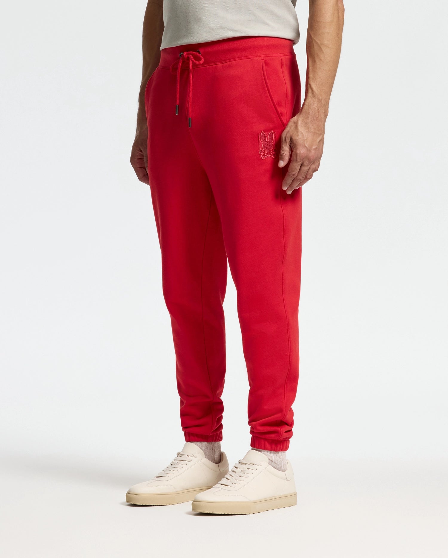 A person wearing the Psycho Bunny MENS PIERCE ESSENTIAL FRENCH TERRY SWEATPANT in bright red, featuring an embroidered logo on the pocket, is paired with a white shirt and light-colored sneakers while standing against a plain background.