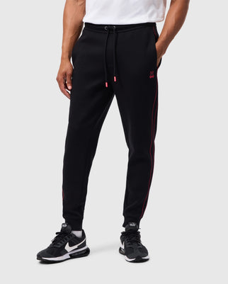 A person is wearing the Psycho Bunny MENS TRAVIS TRACK PANT - B6P143D200, which are high-quality cotton-blend black joggers accented with red along the sides and featuring a small silicone heat transfer bunny logo on the upper thigh. The joggers have a drawstring waistband. They are also sporting black and white sneakers against a plain, light-colored background.