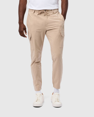 A person wearing Psycho Bunny's MENS LLOYD CARGO JOGGER - B6P159B200 in beige, accompanied by a white T-shirt and white sneakers. The jogger pants feature drawstring ties at the waist, snap fastening pockets, and cargo pockets on the sides. The individual is standing against a plain light gray background.