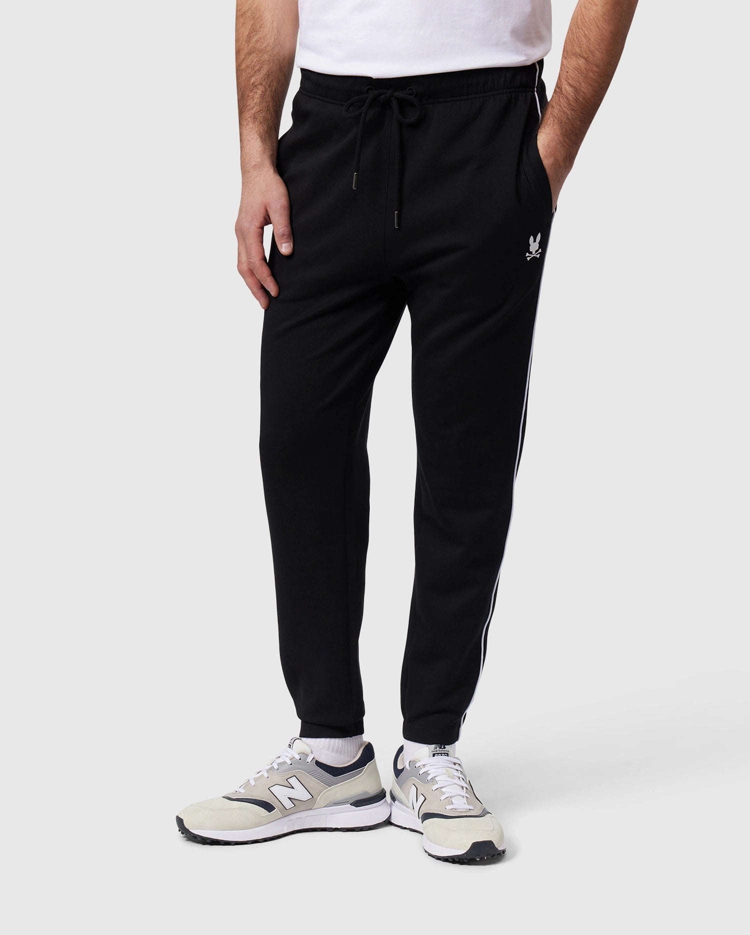 A person wearing a pair of Psycho Bunny MENS LEON REGULAR FIT SWEATPANTS - B6P289B200 in black with a white stripe and logo on the side, paired with light gray and white sneakers. Only the lower half of the person's body is visible.