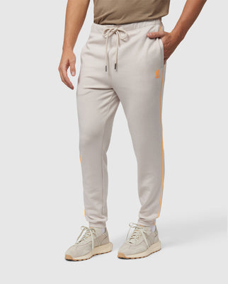 A close-up of a person wearing light grey men's Psycho Bunny Leon Regular Fit sweatpants with an orange stripe and logo, paired with beige sneakers, on a white background.