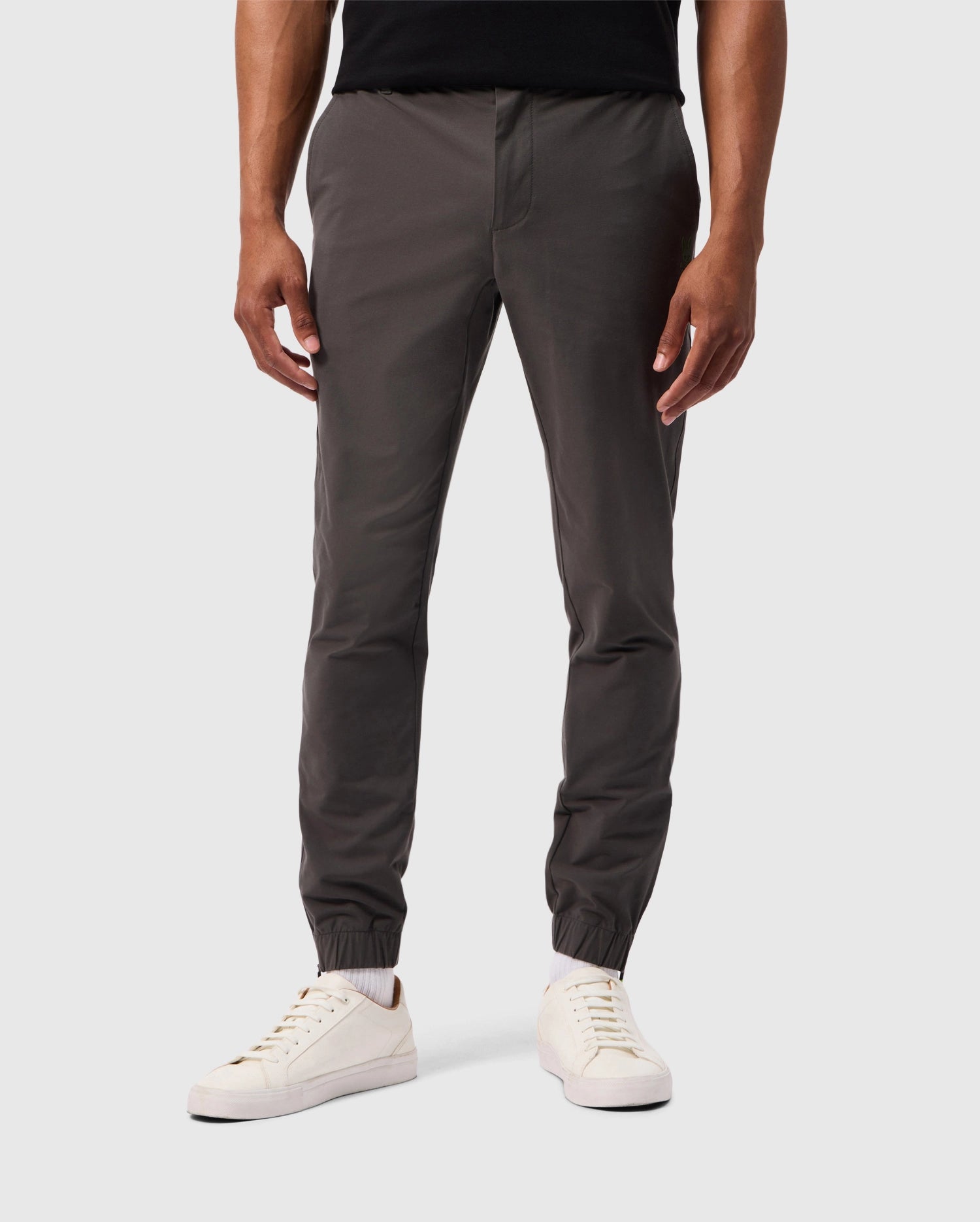 A person in the MENS MADISON COMMUTER PANT - B6P395Z1WB by Psycho Bunny, featuring dark gray jogger pants with elastic cuffs at the ankles and a detachable keychain, stands against a plain gray background in white sneakers. These joggers combine style with practicality. The upper body is not visible.