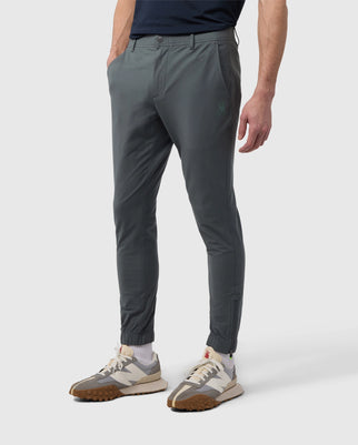 A man stands showing only his lower half, wearing gray Psycho Bunny men's Madison commuter pants with embroidered Bunny logos and stylish sneakers, on a plain white background.