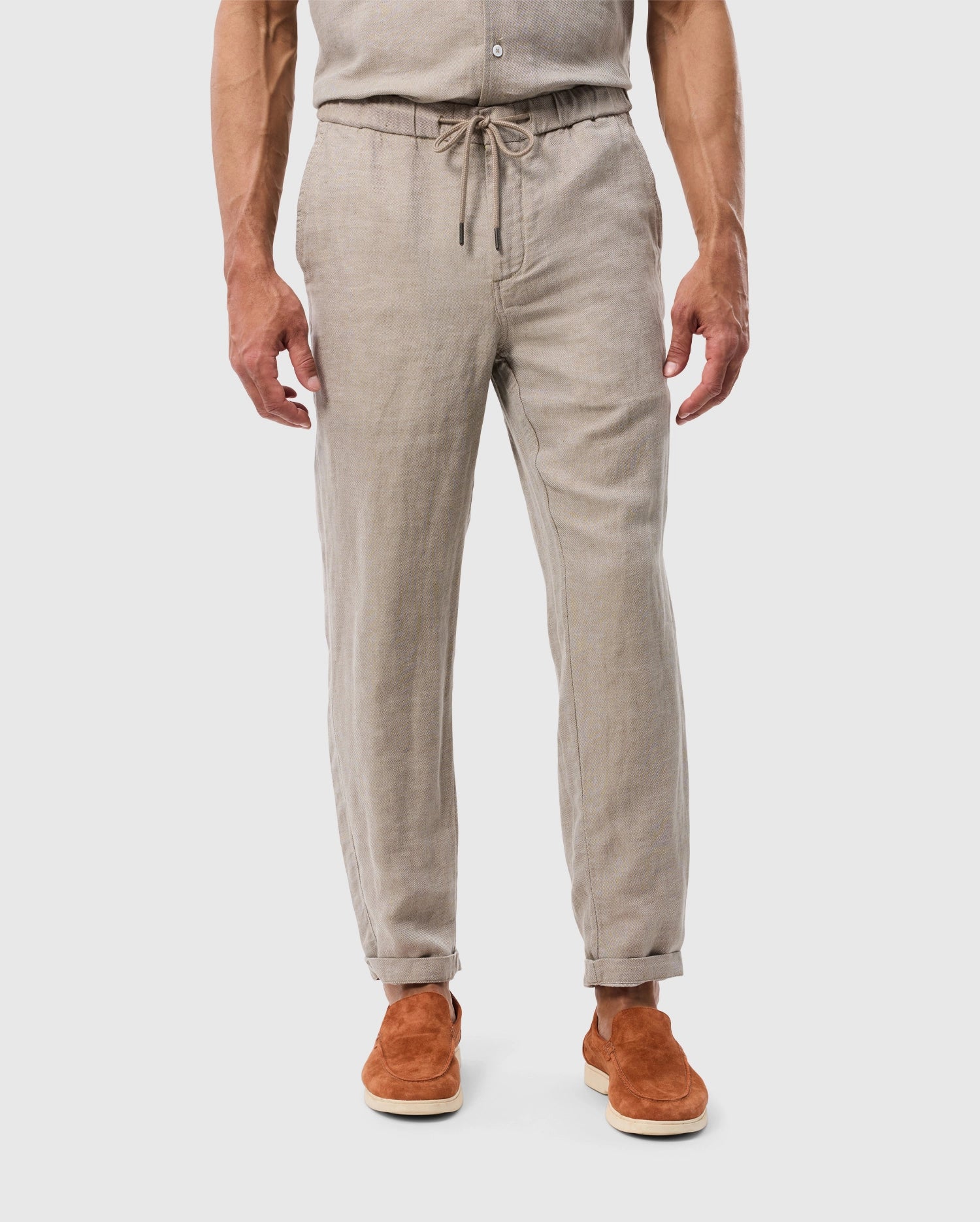 A man stands facing away, showcasing MENS FATE LINEN DRAWSTRING PANTS from Psycho Bunny paired with brown suede shoes. Only the lower half of his body is visible. White background.