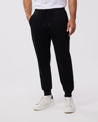 A person is shown from the shoulders down, wearing black MENS OUTLINE SWEATPANT - B6P506ARCN by Psycho Bunny with a drawstring waist and white sneakers. The person has their hands in the pockets of the sweatpants, showcasing the embroidered Bunny logo. The background is plain white.