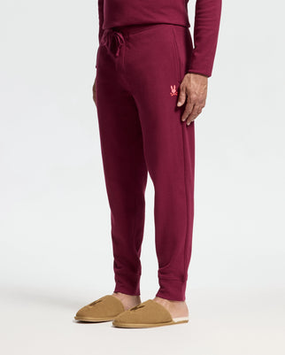 A person wearing Psycho Bunny's MENS VAL WAFFLE LOUNGE PANT - B6P673E200 in burgundy stands against a plain background. These stylish lounge pants feature a small embroidered emblem on the left thigh. The ensemble is completed with a matching burgundy long-sleeve top and brown slippers, perfect for relaxed comfort.