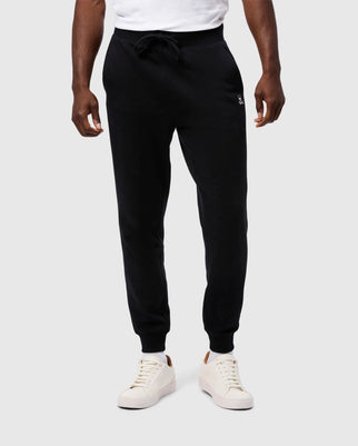 A person is standing against a neutral background wearing Psycho Bunny's MENS LEVY ESSENTIAL FLEECE SWEATPANT (B6P721D200) in black, featuring a drawstring waist. They have on a white T-shirt, white sneakers with tan details, and their hands are relaxed at their sides. Only the lower half of their body is visible.