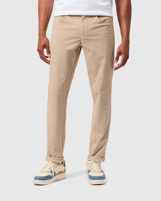 A person is shown from the waist down wearing Psycho Bunny's MENS 10K STEP PANT - B6P788D200, which are light beige five-pocket pants with four-way stretch, paired with a white shirt. The person’s hands are relaxed by their sides. They are also sporting beige and white sneakers with blue accents. The background is plain and light-colored.