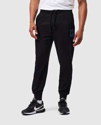 A person is shown from the shoulders down, wearing a pair of black Psycho Bunny MENS WALTER LIGHTWEIGHT SWEATPANTS (B6P860D200) featuring a blue bunny logo on the upper left thigh. The person is also wearing black and white athletic shoes. The background is plain white.