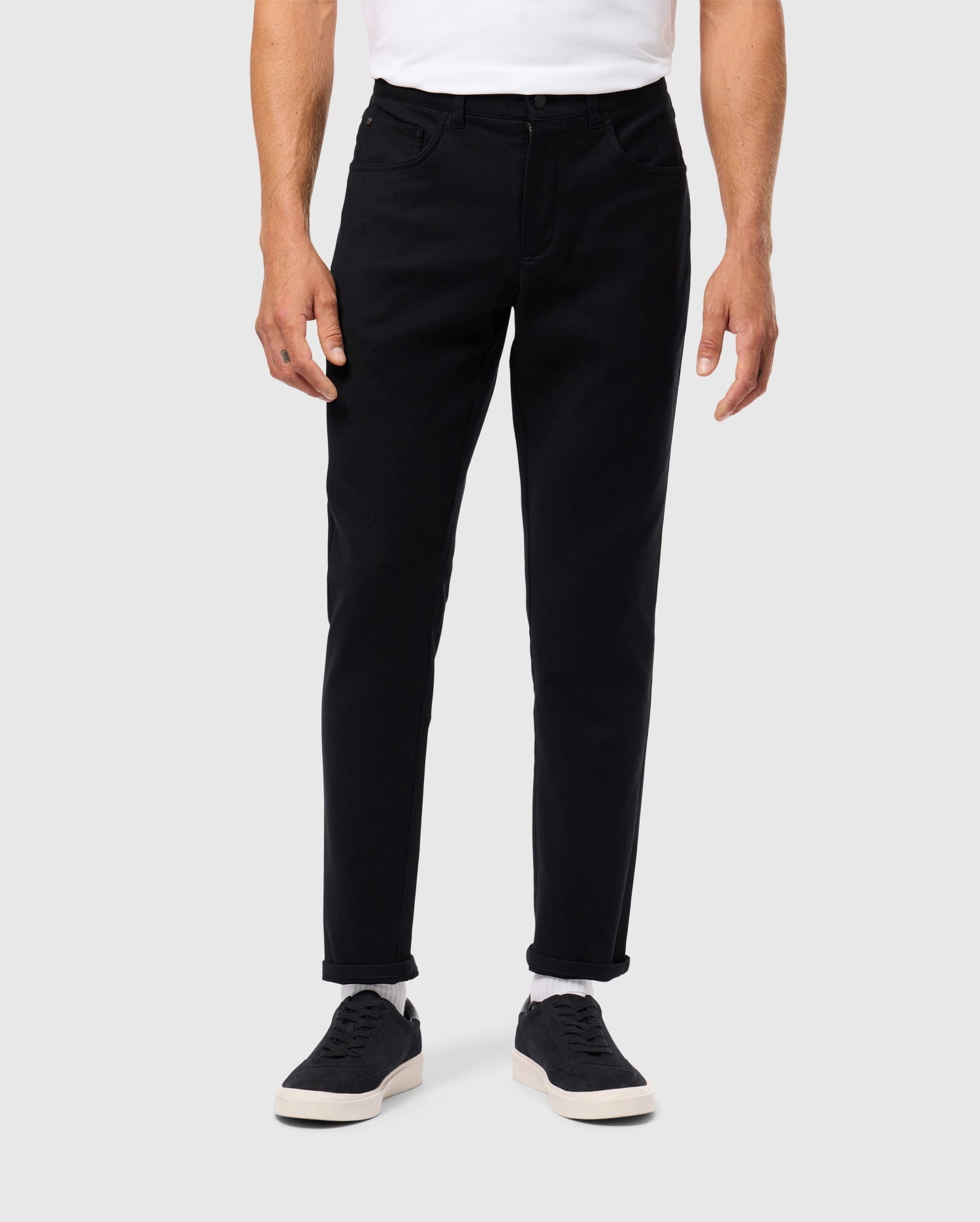 A person is wearing Psycho Bunny's MENS HAYS TWILL PANT - B6P894D200 in black, featuring a cotton-blend twill fabric with a slim fit, paired with a white shirt and black sneakers accented by white soles for a casual touch. The image is framed from the shoulders down against a plain backdrop.