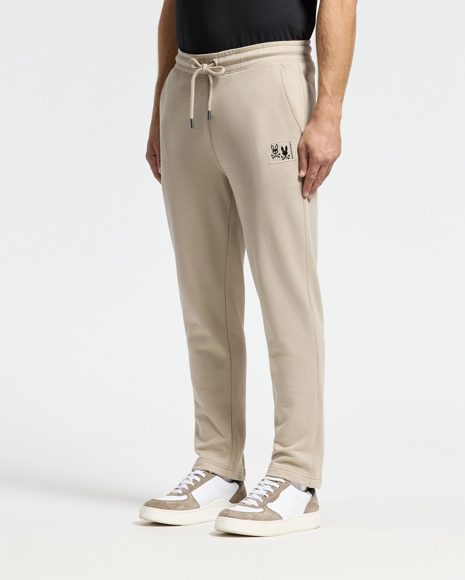 A person is dressed in Psycho Bunny's MENS MONROE TWILL PATCH FLEECE SWEATPANT in beige, featuring a subtle logo on the pocket, and paired with white and beige sneakers. The plain white background accentuates the casual ensemble.