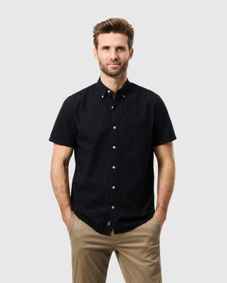 A man with short, brown hair and a trimmed beard is standing against a plain white background. He is wearing a short-sleeved, button-up black Psycho Bunny MENS ALTON SEERSUCKER SHIRT - B6Q147C200 and beige pants, with his hands casually placed in his pockets. He is looking towards the camera and smiling subtly.