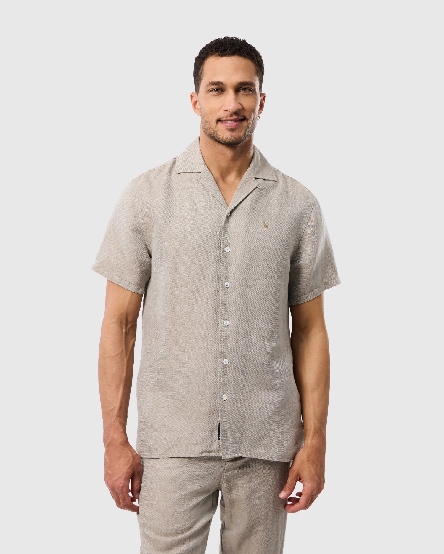 Man in a Psycho Bunny MENS WINDCREST LINEN BLEND SHORT SLEEVE SHIRT with an embroidered logo and matching pants standing against a white background, smiling slightly at the camera. He has a casual, well-groomed appearance.