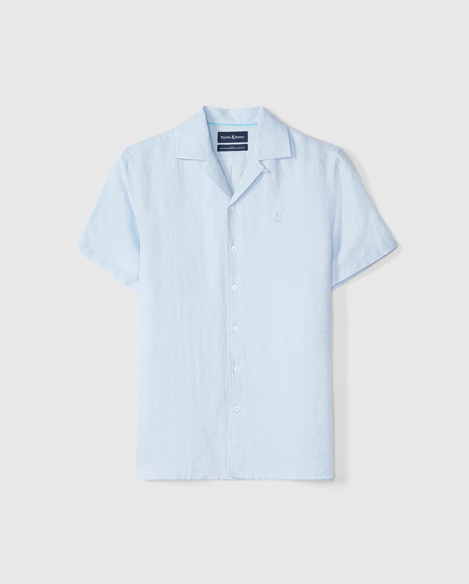 A light blue Psycho Bunny MENS WINDCREST LINEN SHORT SLEEVE SHIRT - B6Q148C200 is displayed against a plain white background. It features a small, subtle embroidered logo on the left chest area and a navy tag at the inside collar.