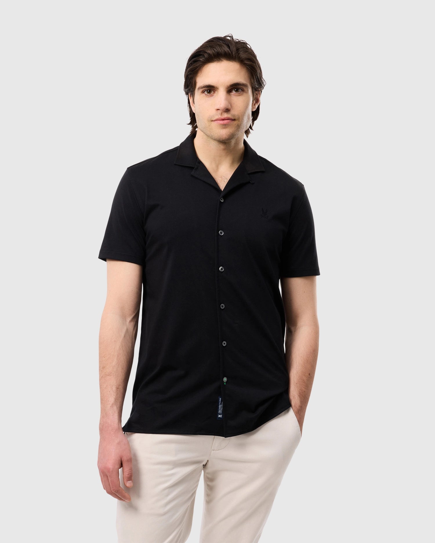 A man with dark hair wearing a black Psycho Bunny MENS KELLER CAMP COLLAR SHORT SLEEVE SHIRT - B6Q448C200 made from a Pima cotton-blend and white pants stands against a plain white background. His right hand is in his pocket while his left arm rests by his side. He has a neutral expression on his face.