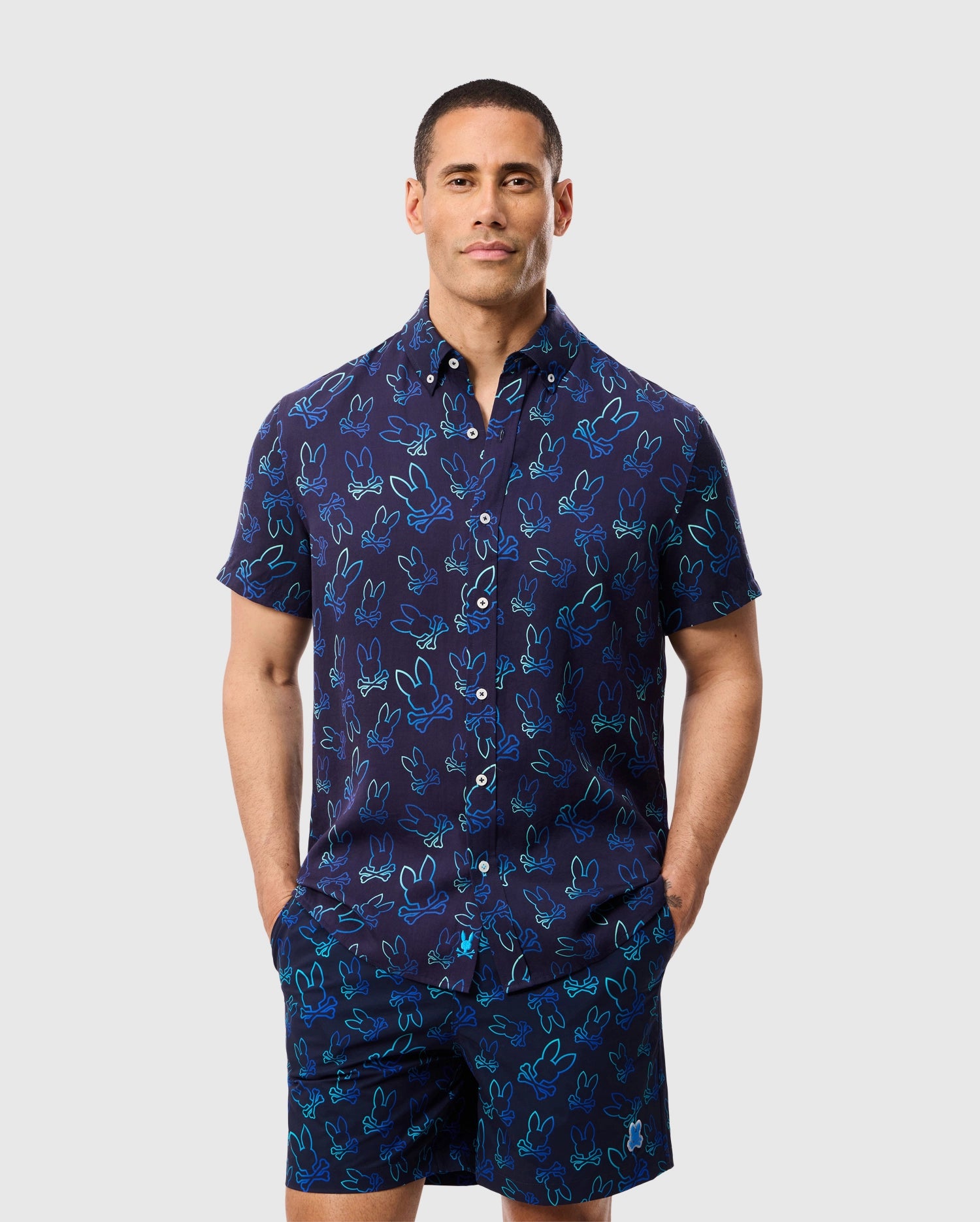 A man stands against a plain background wearing a matching short-sleeve shirt and shorts set. The outfit is dark blue with a playful Bunny pattern in light blue and turquoise. He has short hair and his hands are in his pockets, effortlessly showcasing the Psycho Bunny MENS SHELDON ALL OVER PRINT SHIRT - B6Q574C200.