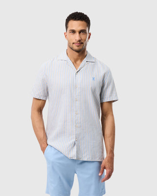 A man with short curly hair is wearing a Psycho Bunny MENS JUSTIN LINEN CAMP SHIRT - B6Q585C200 with light blue and white vertical stripes. The mens linen shirt has a small logo on the left chest. He is also wearing light blue shorts and standing against a plain white background.