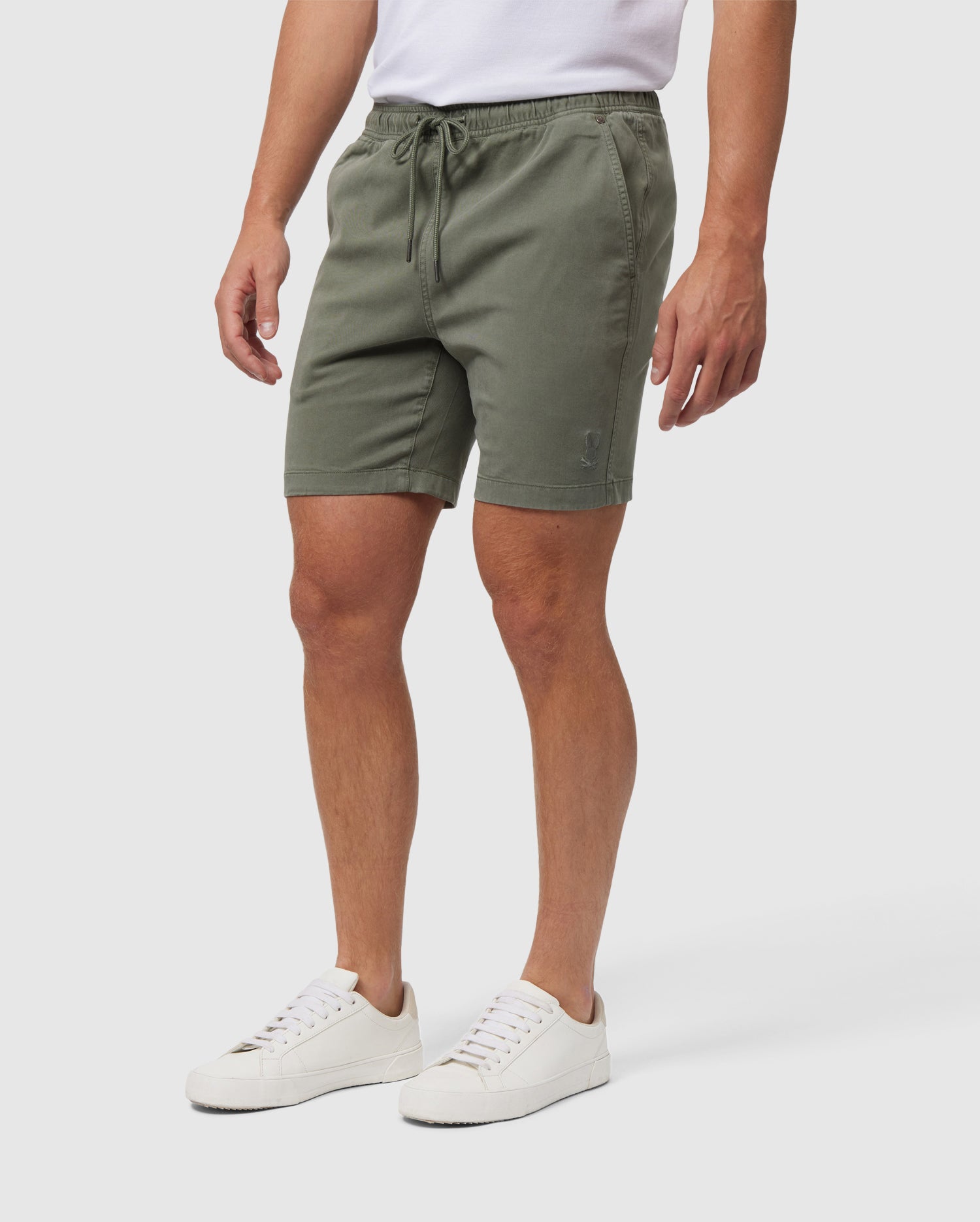 A man wearing Psycho Bunny's MENS WILLIS STRETCH TENCEL SHORT in olive green and white sneakers stands against a white background, with the focus on his lower body from waist to feet.