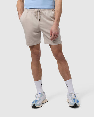 A man wearing light beige Psycho Bunny Men's Willis Stretch Tencel Shorts, white socks with a logo near the ankle, and blue and white sneakers stands against a white background, focusing on his legs and midsection.