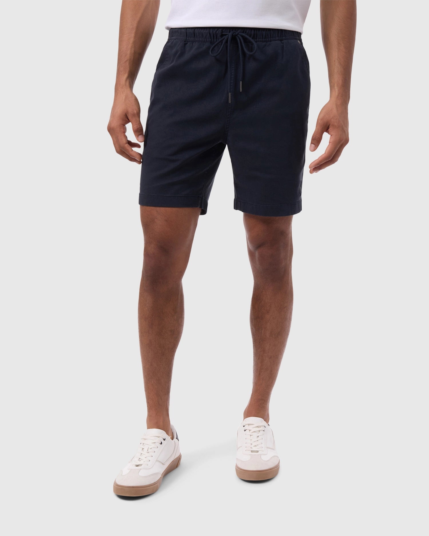 A person wearing Psycho Bunny's MENS WILLIS STRETCH TENCEL SHORT in dark blue and white sneakers stands against a white background, showcasing the outfit clearly. Only the lower half from the waist down is visible.