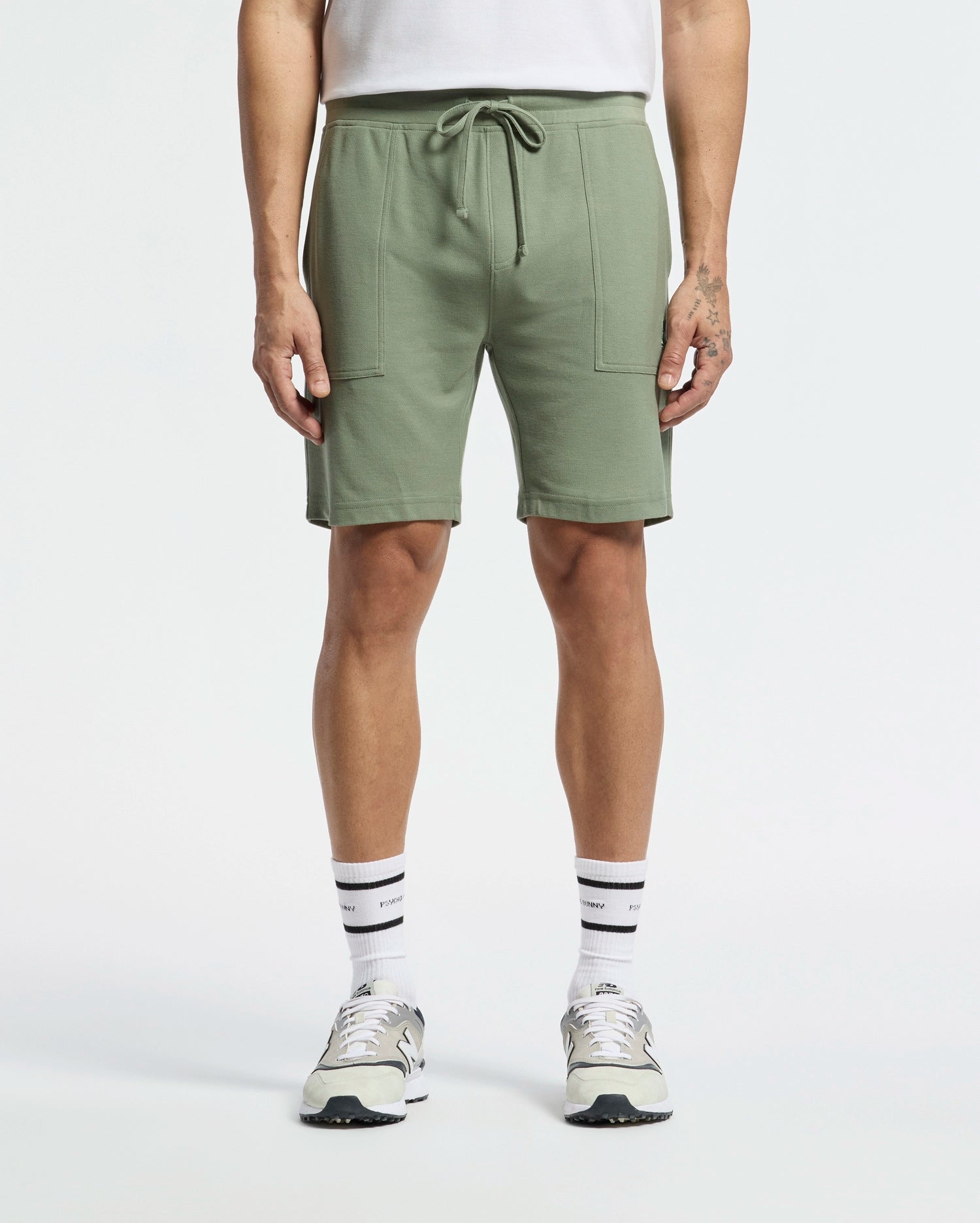 Sporting a casual-chic look, the person wears Psycho Bunny's MENS TAYLOR LIGHT PIQUE SWEATSHORT - B6R344F200 in light green with a drawstring and large front pockets, paired with white socks, gray and white sneakers, and a tucked-in white shirt against a plain white background.