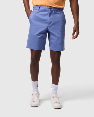 A person is standing wearing blue **Psycho Bunny MENS YORK CHINO SHORT - B6R357B200**, a light orange top, white socks, and white sneakers. The image focuses on the person's legs and midsection, with their hands resting in their pockets. The background is plain white.