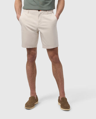 A man in a light green shirt and Psycho Bunny beige MEN'S YORK CHINO SHORT - B6R357B200 stands with his back to the camera, wearing brown suede shoes, isolated on a white background.