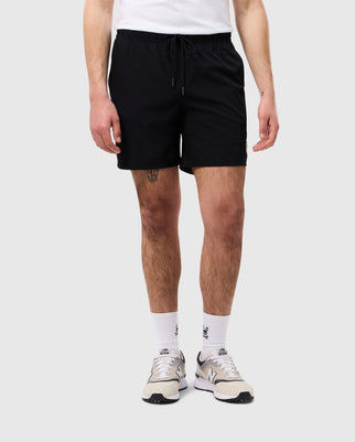 A person is standing against a plain background, wearing a white t-shirt, Psycho Bunny MENS SONTERRA STRIPE SPORT SHORT - B6R464C200 made of breathable fabric, white socks, and beige and grey sneakers. The person's left thigh has a small tattoo. Only the lower half of their body is visible.