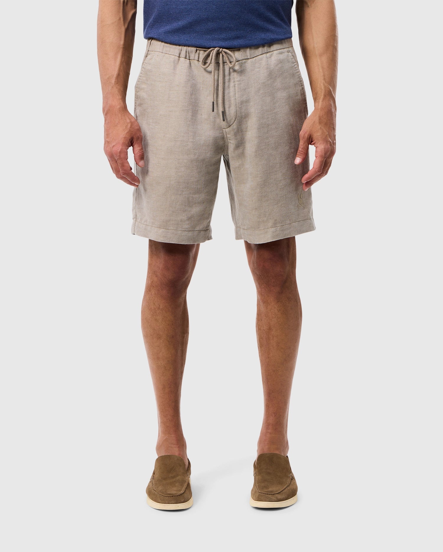 A man stands wearing light gray Psycho Bunny mens Windcrest linen shorts and brown suede shoes, isolated on a white background, showing only from the waist down.