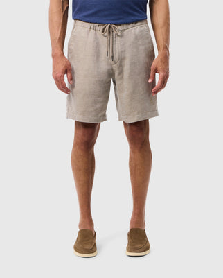 A man stands wearing light gray Psycho Bunny mens Windcrest linen shorts and brown suede shoes, isolated on a white background, showing only from the waist down.