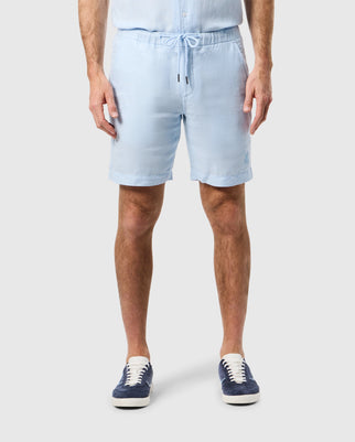 A man wearing light blue Psycho Bunny mens windcrest linen shorts and blue sneakers stands against a white background, showing only from the waist down.