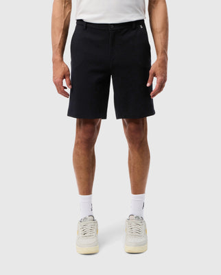 A man stands facing forward wearing a plain black pair of Psycho Bunny MENS SHIRO KNIT HONEYCOMB shorts, white socks, and beige sneakers. Only his lower body from the waist down is visible.