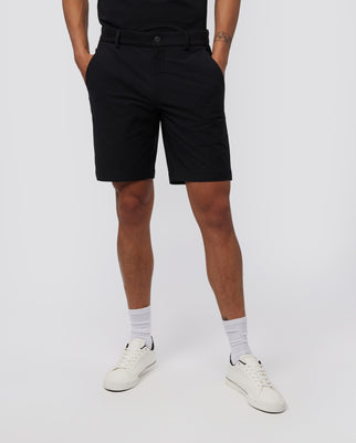 A person wearing black knee-length Psycho Bunny MENS GABLE REGULAR FIT SPORT SHORT - B6R761ARCN, a black t-shirt, white socks, and white sneakers stands against a plain white background. Their hands are in their pockets, showcasing the hidden zip pocket feature of their durable stretch cotton-nylon shorts. Only the lower half of their body is visible.