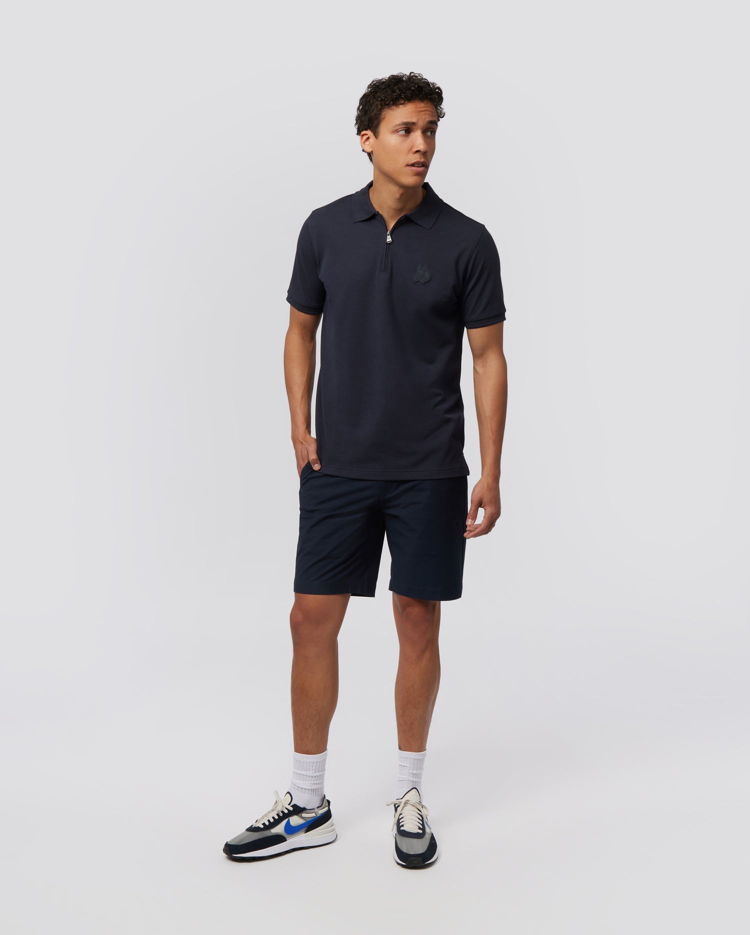 A person stands against a plain white background wearing a navy blue polo shirt and Psycho Bunny MENS GABLE REGULAR FIT SPORT SHORT - B6R761ARCN made from durable stretch cotton-nylon. They have short, curly hair and are dressed casually with white socks and white sneakers featuring blue and black accents. The shorts feature a hidden zip pocket for convenience.