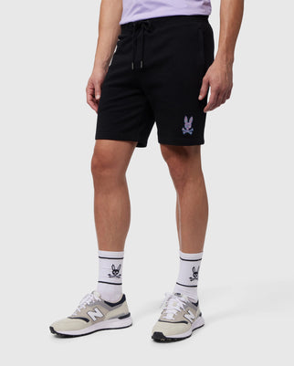 A person is wearing a black pair of Psycho Bunny MENS SANTA CRUZ SWEATSHORT - B6R922A2FT featuring a small graphic of a bunny on the left leg. They are also wearing white socks with the same bunny graphic, and white and gray New Balance sneakers. The background is plain white.