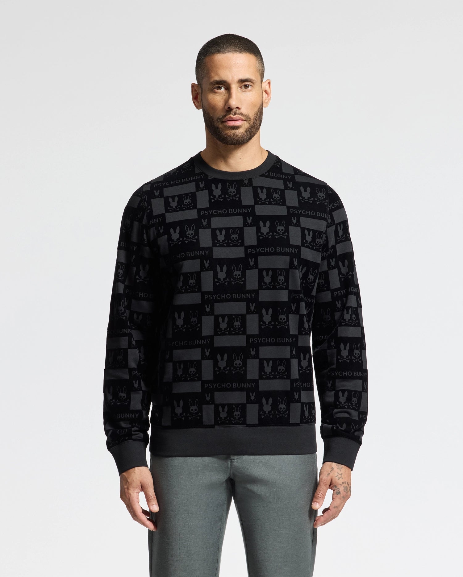 A man with short hair and a beard is wearing the MENS ROMEO LIGHTWEIGHT SWEATSHIRT - B6S129E200 by Psycho Bunny, featuring a sleek black design with a repeating pattern of checkerboard designs and 