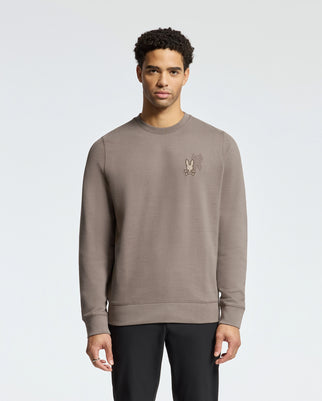 A man with short curly hair stands against a plain background wearing Psycho Bunny's stylish light brown MENS PIERCE PEACHED TERRY CREW, B6S278E200. The sweatshirt features embroidery and a small cartoon character on the left chest, paired with black pants.