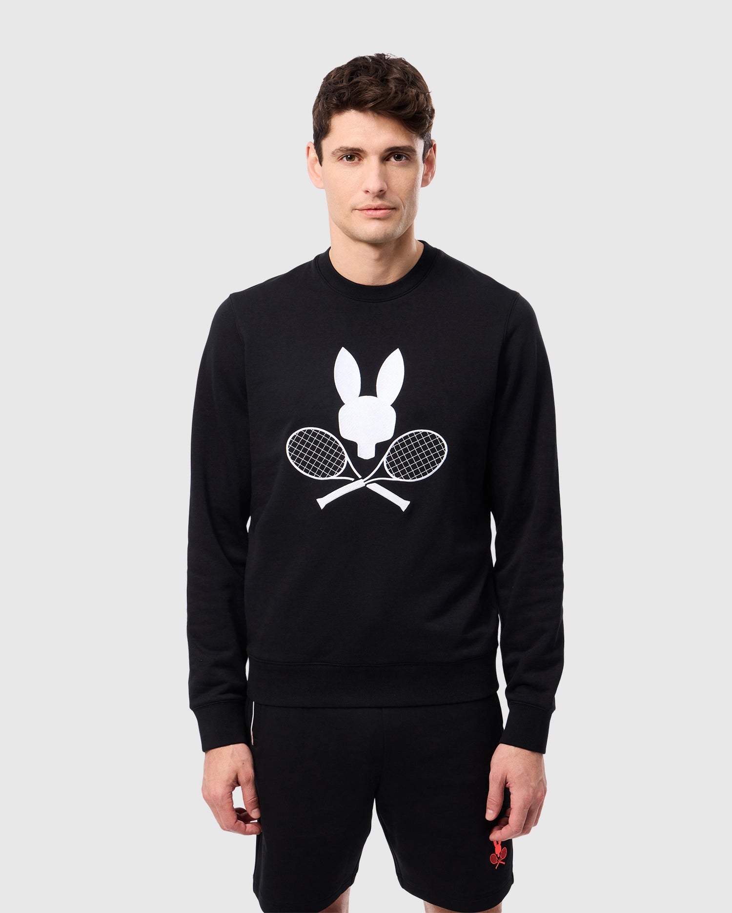 A man displays a tennis-inspired Psycho Bunny MENS COURTSIDE SWEATSHIRT - B6S688C200, crafted from a breathable cotton-blend in black and featuring a white graphic of a bunny head and crossed tennis rackets. He pairs it with black shorts, standing against a plain light gray backdrop, exuding sporty elegance.