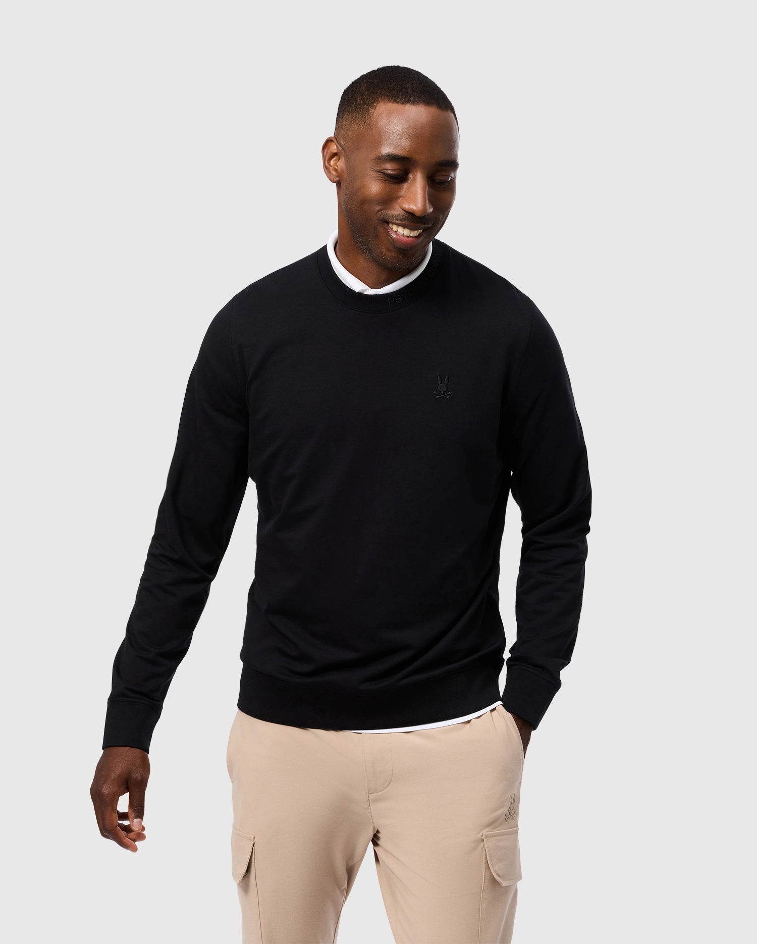 A person is smiling while wearing a Psycho Bunny MENS DUKE EMBROIDERED SWEATSHIRT - B6S706D200 in black paired with beige pants against a plain light gray background.