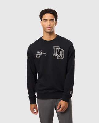 A young man stands against a plain background, wearing the Psycho Bunny MENS ZACK VARSITY SWEATSHIRT - B6S880D200. The black long-sleeve sweatshirt features white chenille embroidery patches, including a large "DB" logo with a rabbit graphic on the left chest and the words "Psycho Bunny" with a smaller rabbit on the right chest.