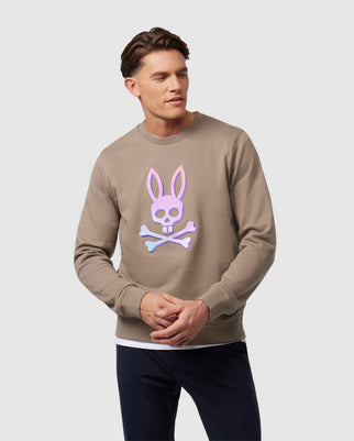 A person is wearing a Psycho Bunny men's Bloomington ombre embroidered crew sweatshirt (B6S924A2FT) with a pink bunny skull and crossbones design on the front. They are standing against a plain white background, clasping their hands and wearing dark pants.