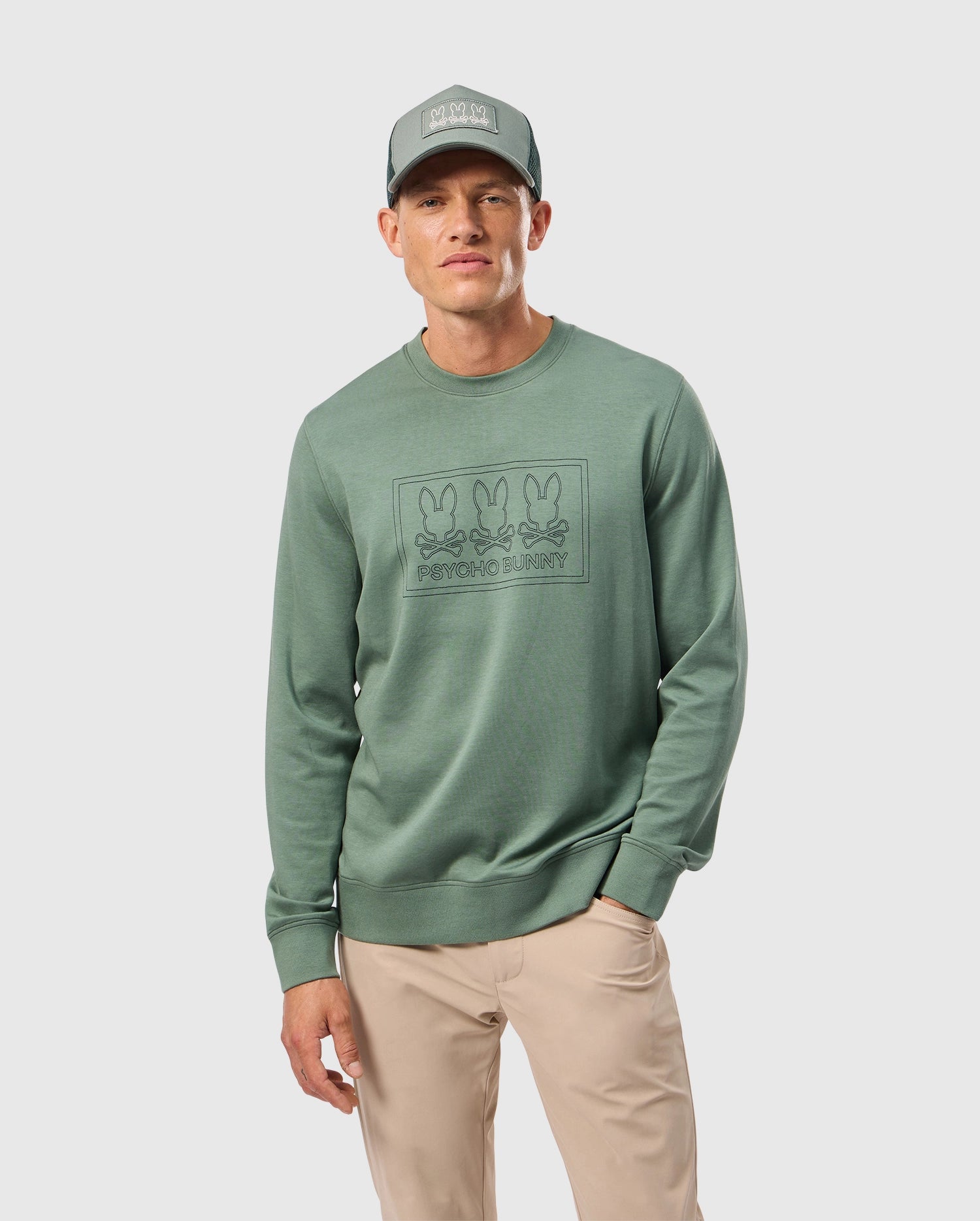 Dressed in the MENS TACOMA SWEATSHIRT by Psycho Bunny, a person stands against a plain white background. The green sweatshirt features a graphic of three bunnies and 