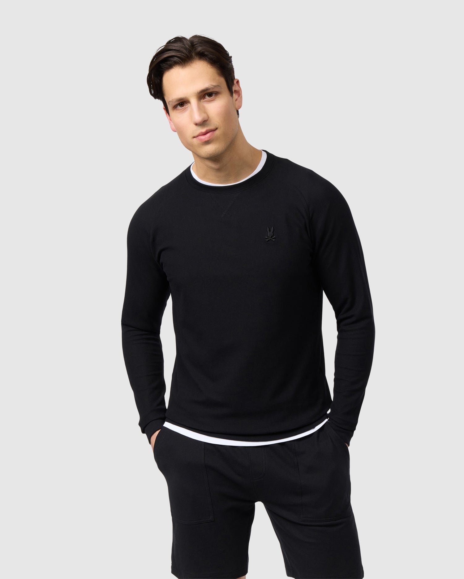 A man wearing a Psycho Bunny MENS STANFORD LONG SLEEVE PIQUE TEE - B6T341B200 and shorts stands against a light gray background, looking directly at the camera with a neutral expression.